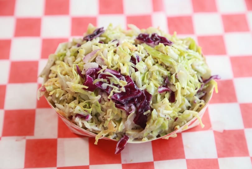 Creamy coleslaw with green and purple cabbage in a paper boat on a checkered background.