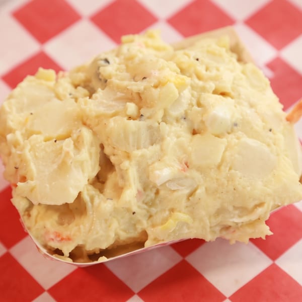 Creamy potato salad with specks of pepper and vegetables in a paper boat on a checkered background.