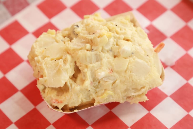 Creamy potato salad with specks of pepper and vegetables in a paper boat on a checkered background.